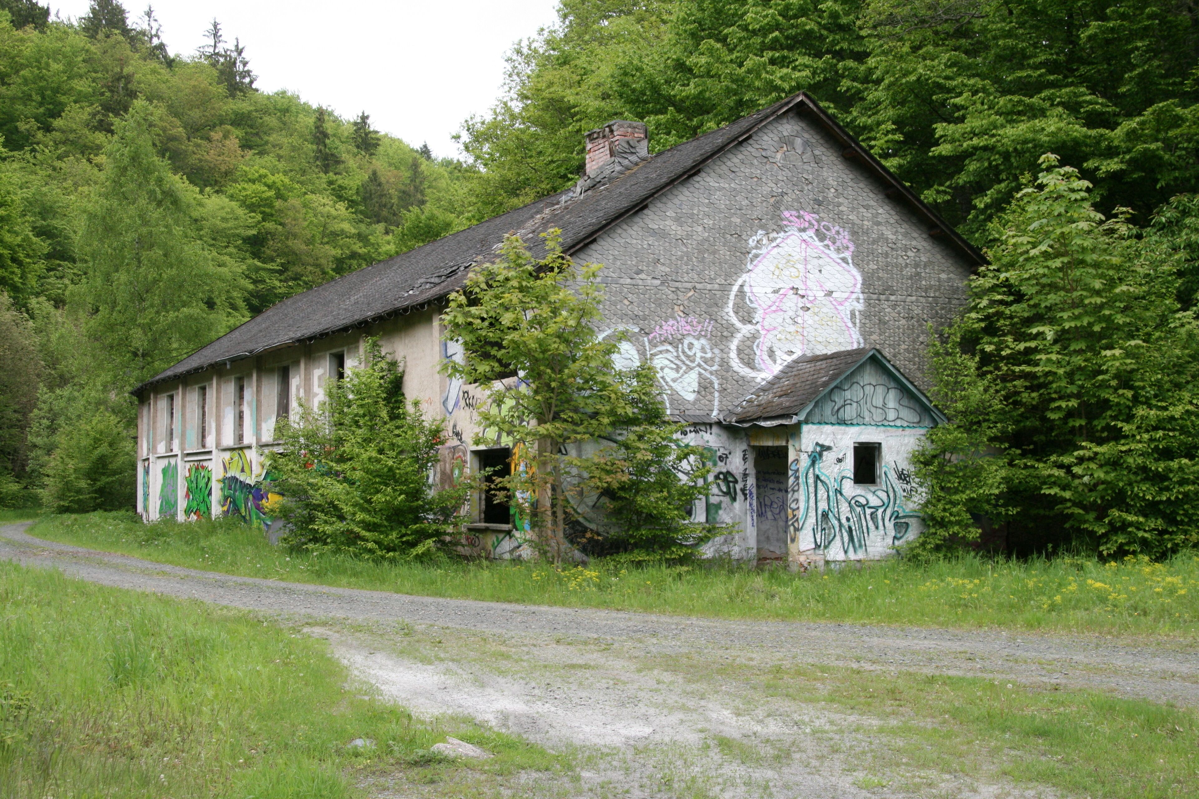 An abandoned building with graffiti, surrounded by trees and a gravel path.
