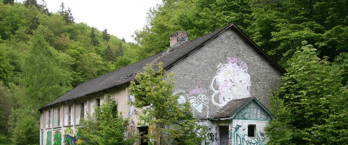 An abandoned building with graffiti, surrounded by trees and a gravel path.