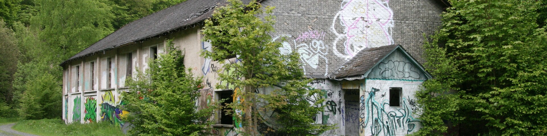 An abandoned building with graffiti, surrounded by trees and a gravel path.