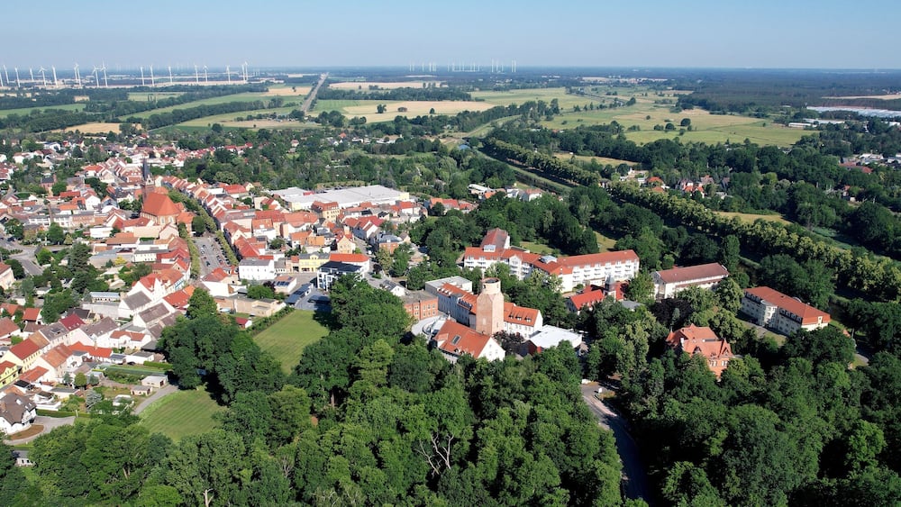An aerial view of a town with red-roofed buildings, green fields, and wind turbines in the distance.