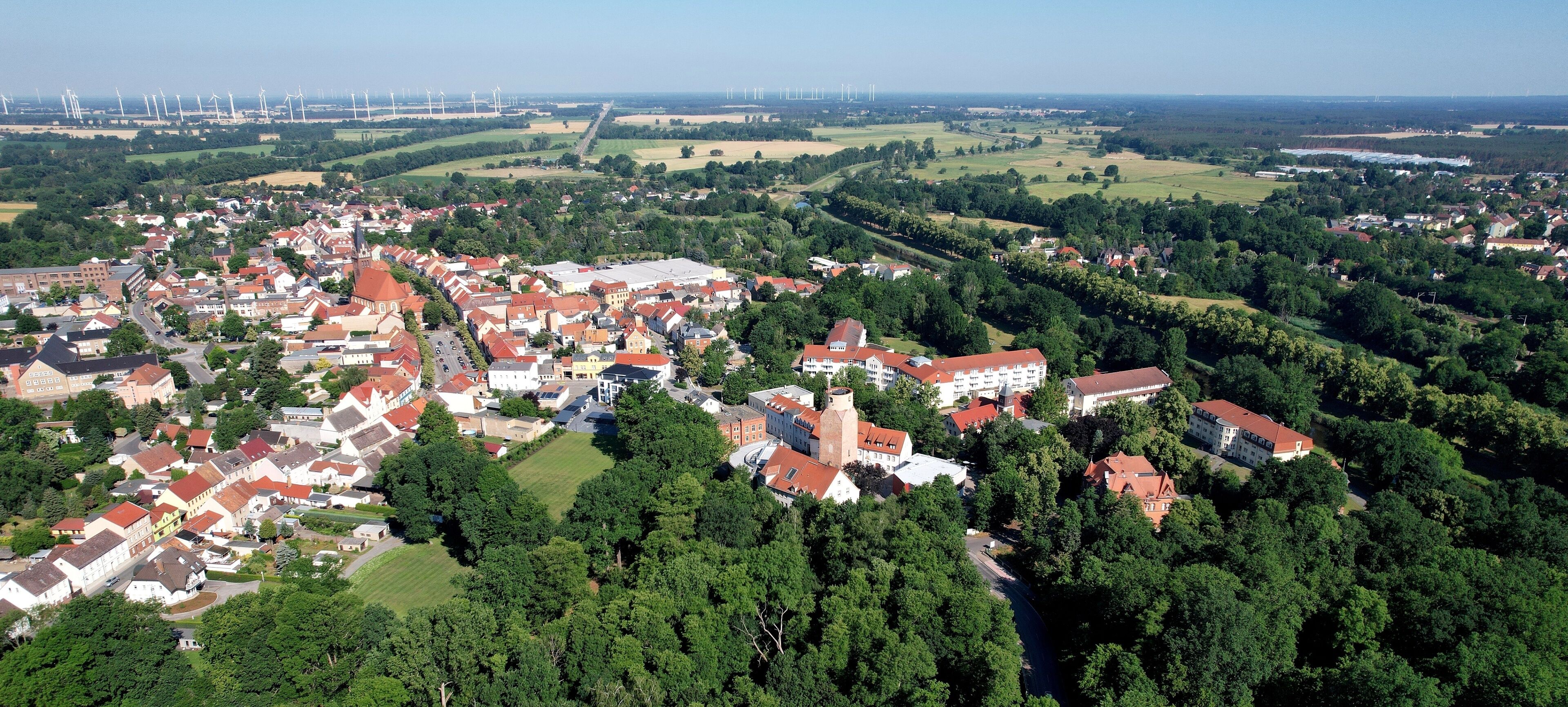 An aerial view of a town with red-roofed buildings, green fields, and wind turbines in the distance.