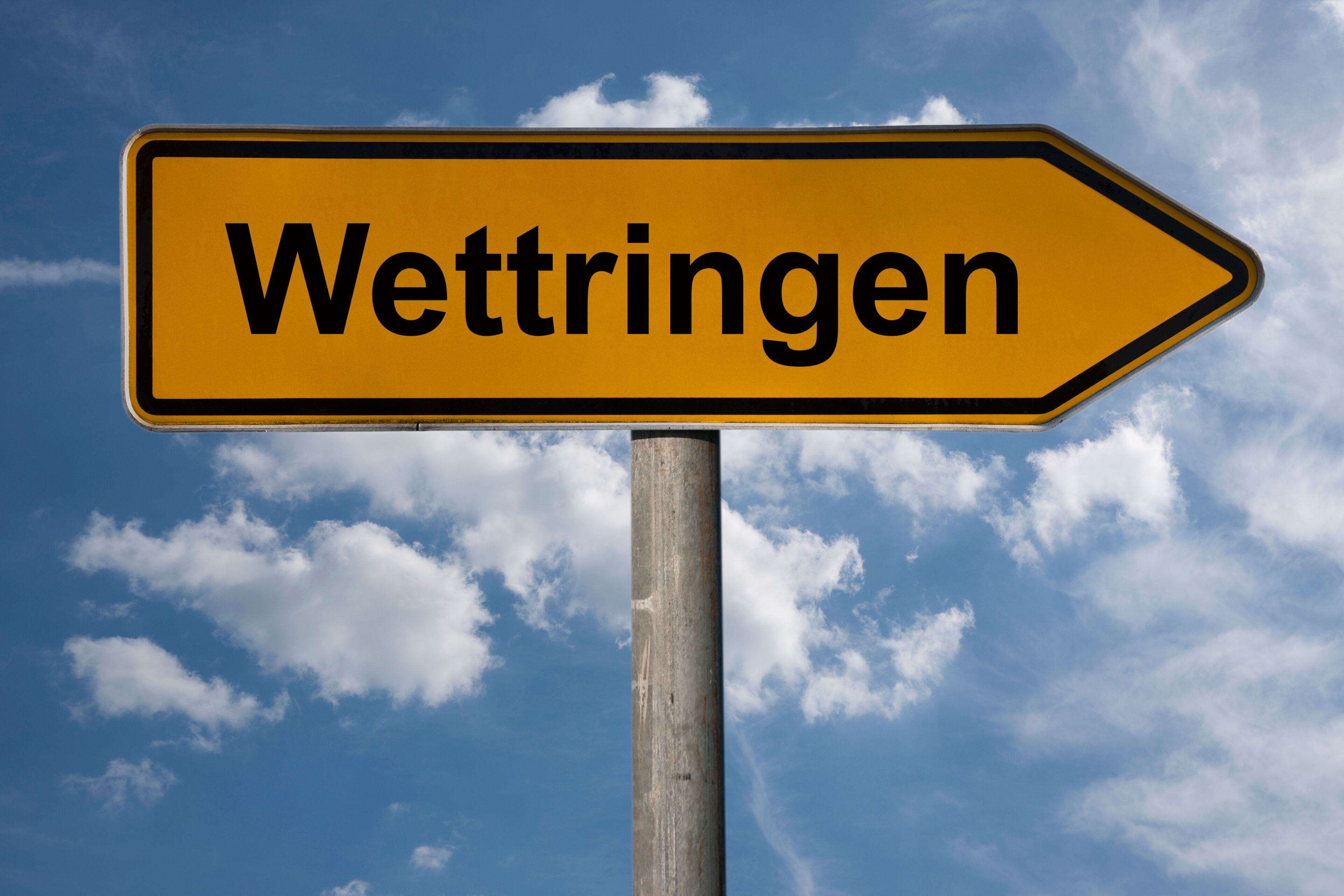 A directional sign with the word "Wettringen" against a blue sky with clouds.