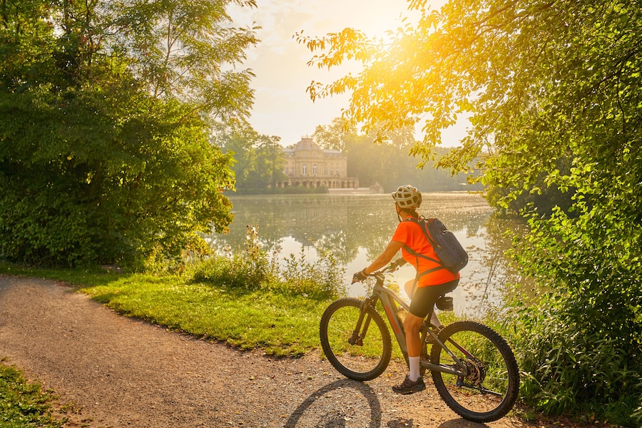 A cyclist on a gravel path beside a calm lake, with lush greenery and a historic building in the background.