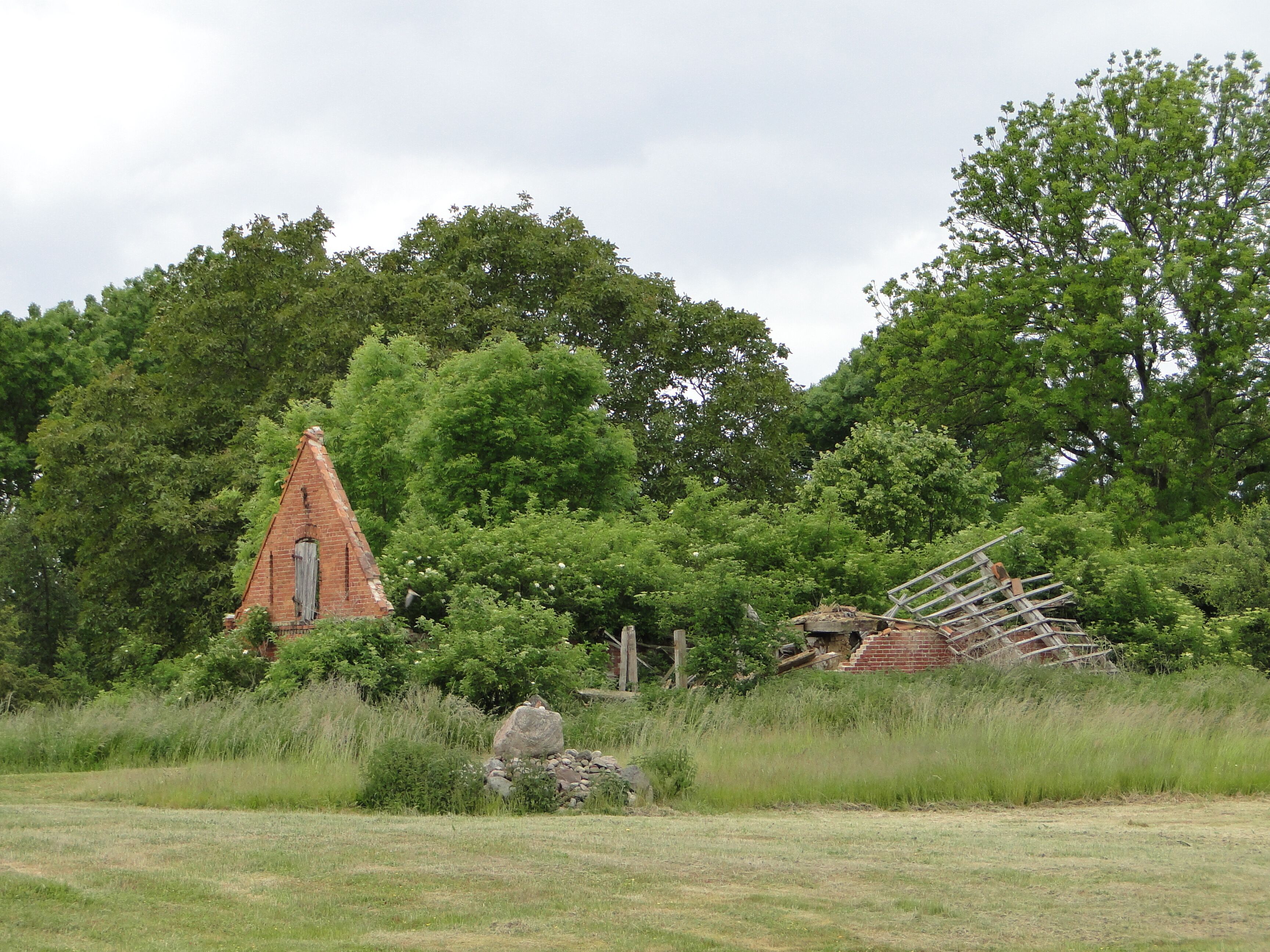 A dilapidated brick structure with a pointed roof, surrounded by lush greenery and trees.