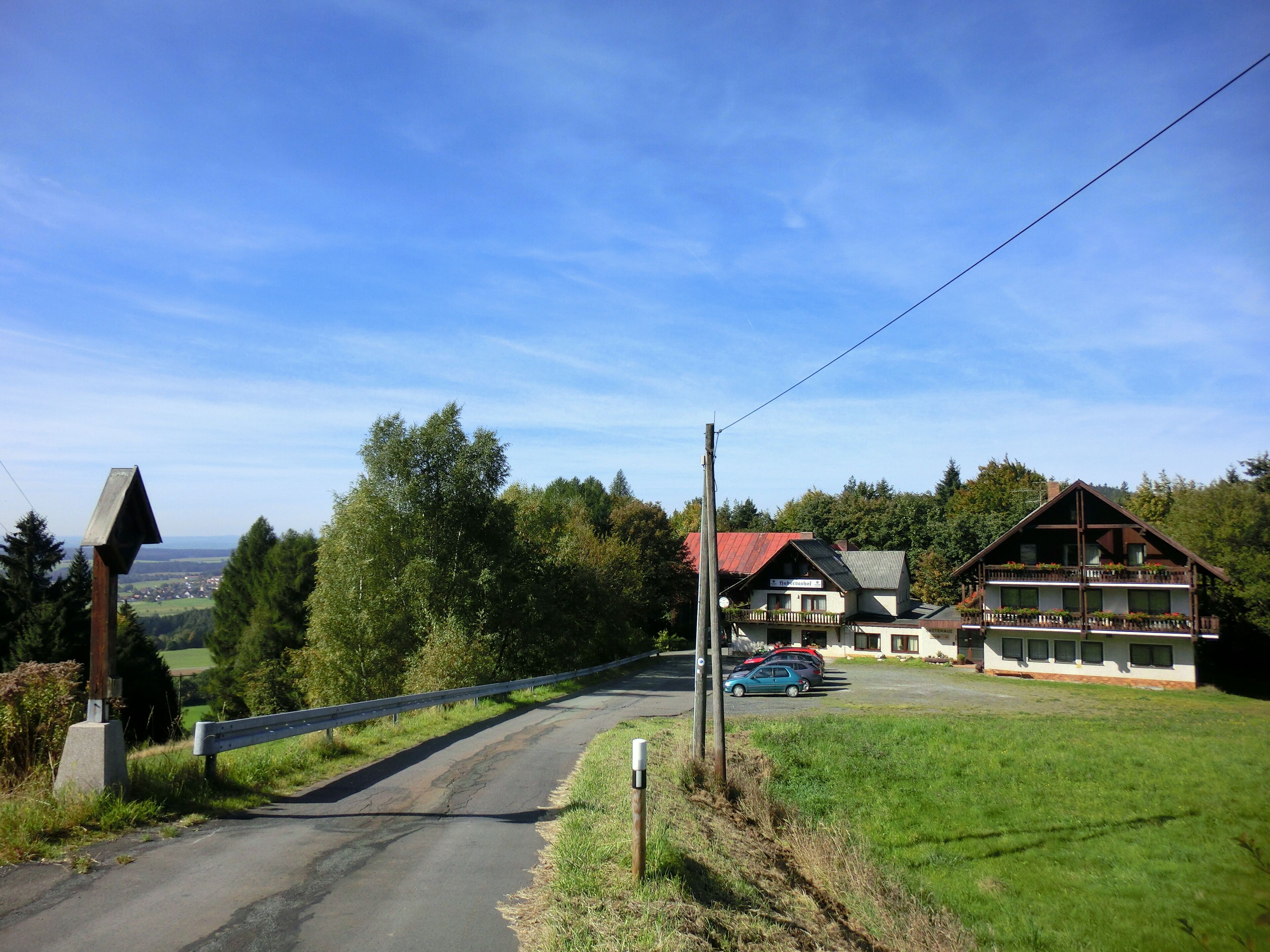 A rural road with a wooden guardrail, a blue car parked near a building with a red roof, and a view of a valley in the distance.