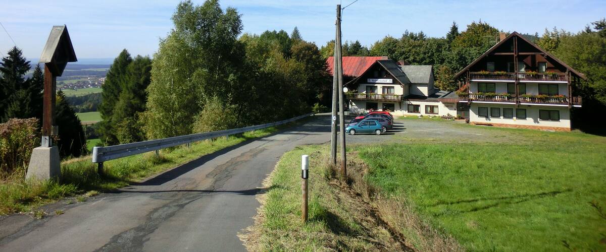 A rural road with a wooden guardrail, a blue car parked near a building with a red roof, and a view of a valley in the distance.