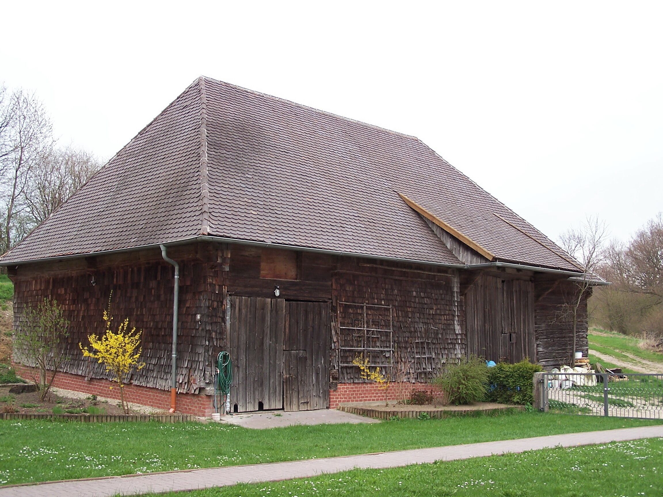 A rustic wooden barn with a sloped roof, surrounded by a grassy area and a small fence.