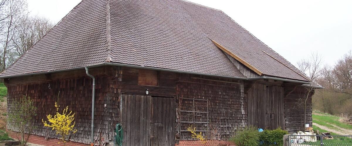 A rustic wooden barn with a sloped roof, surrounded by a grassy area and a small fence.
