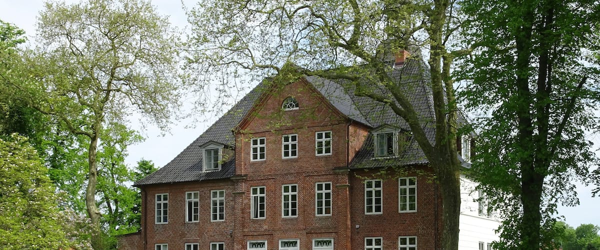 A large brick house with a steep roof, surrounded by lush green grass and trees.
