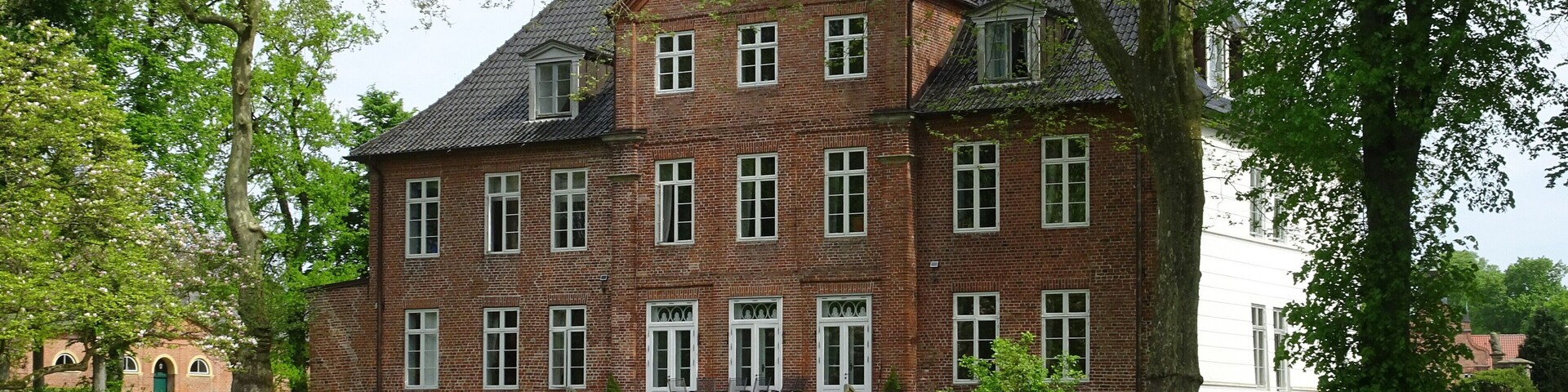 A large brick house with a steep roof, surrounded by lush green grass and trees.