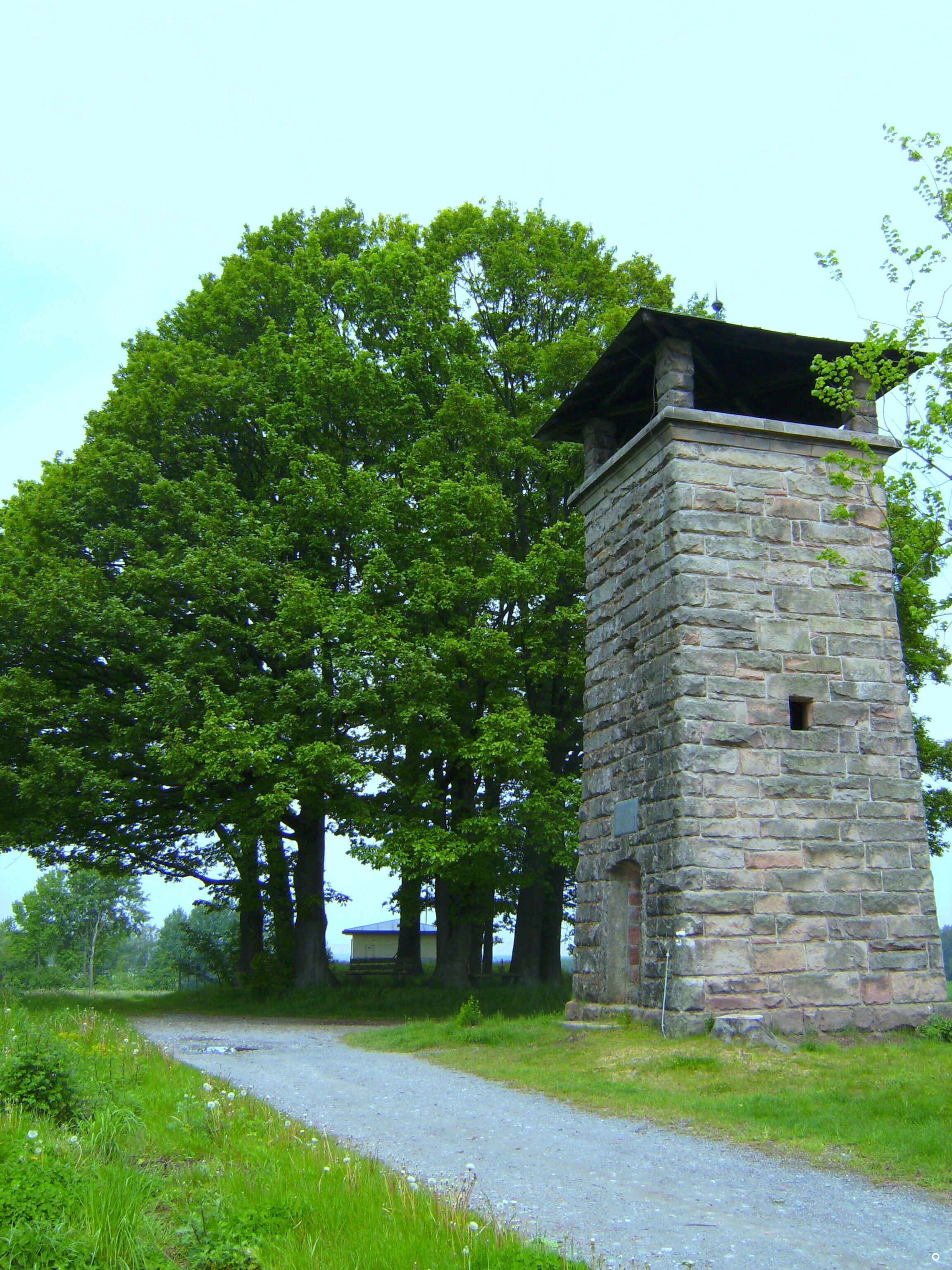 A stone tower with a metal roof, surrounded by lush green trees and a gravel path.