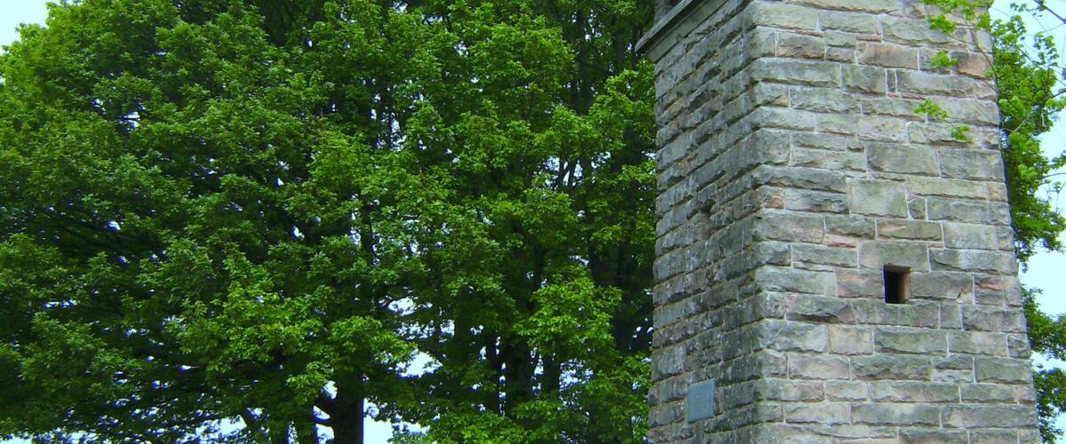 A stone tower with a metal roof, surrounded by lush green trees and a gravel path.