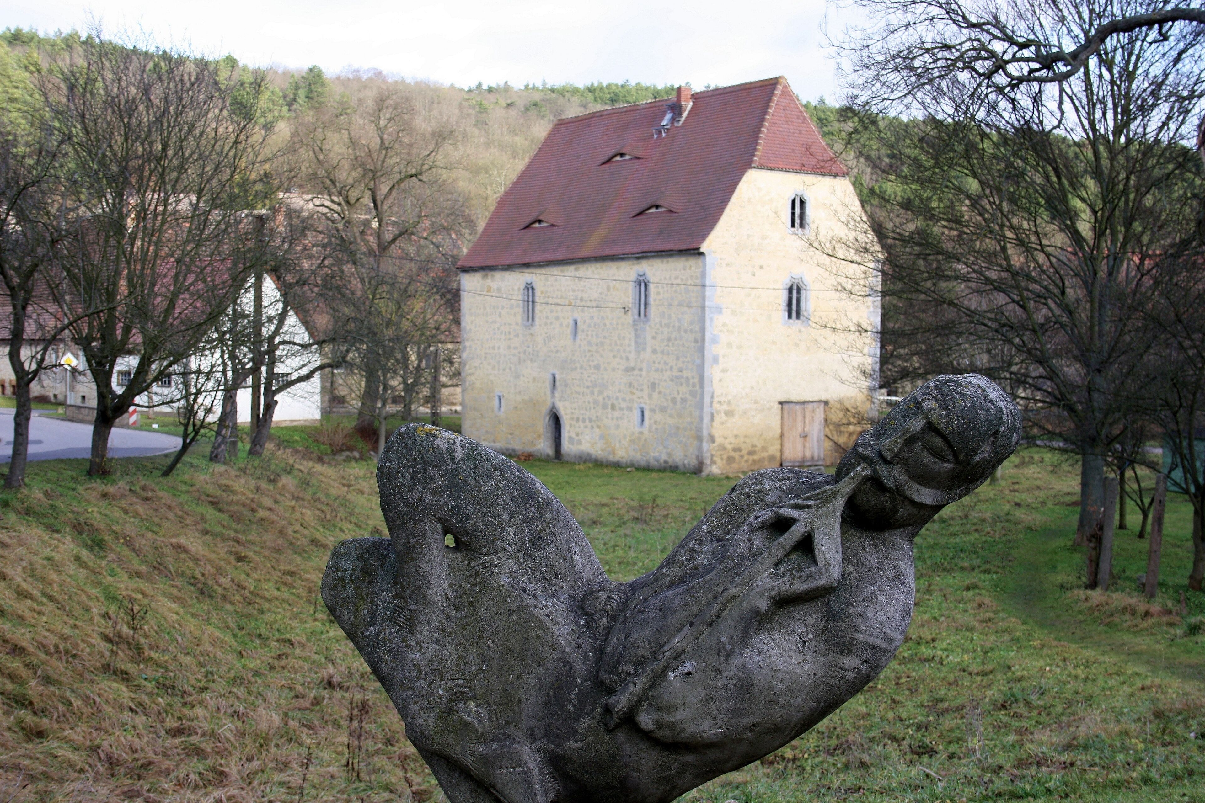 A stone sculpture of a hand holding a heart, a stone building with a red roof, and a grassy hill with trees in the background.
