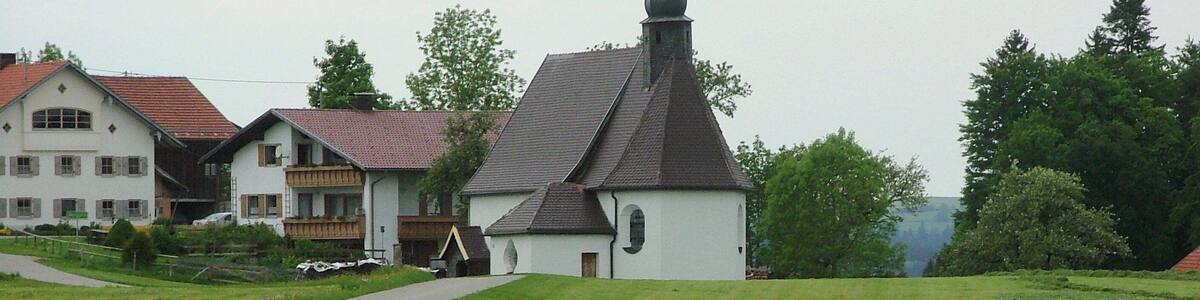 Une église dotée d’un toit à forte pente et d’un petit clocher, entourée de champs verdoyants et de quelques immeubles d’habitation.