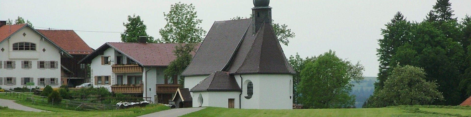 A church with a steeply pitched roof and a small tower, surrounded by lush green fields and a few residential buildings.