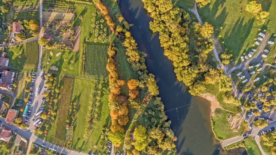 An aerial view of a riverside area with fields, trees, and RVs parked along the road.