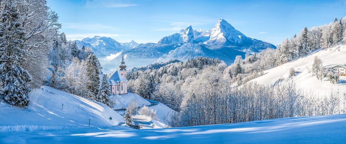 A snow-covered mountain landscape with a church, a house, and a clear blue sky.