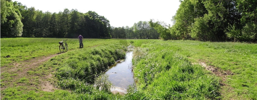 A narrow water channel in a grassy field with a bicycle and a person standing in the distance.