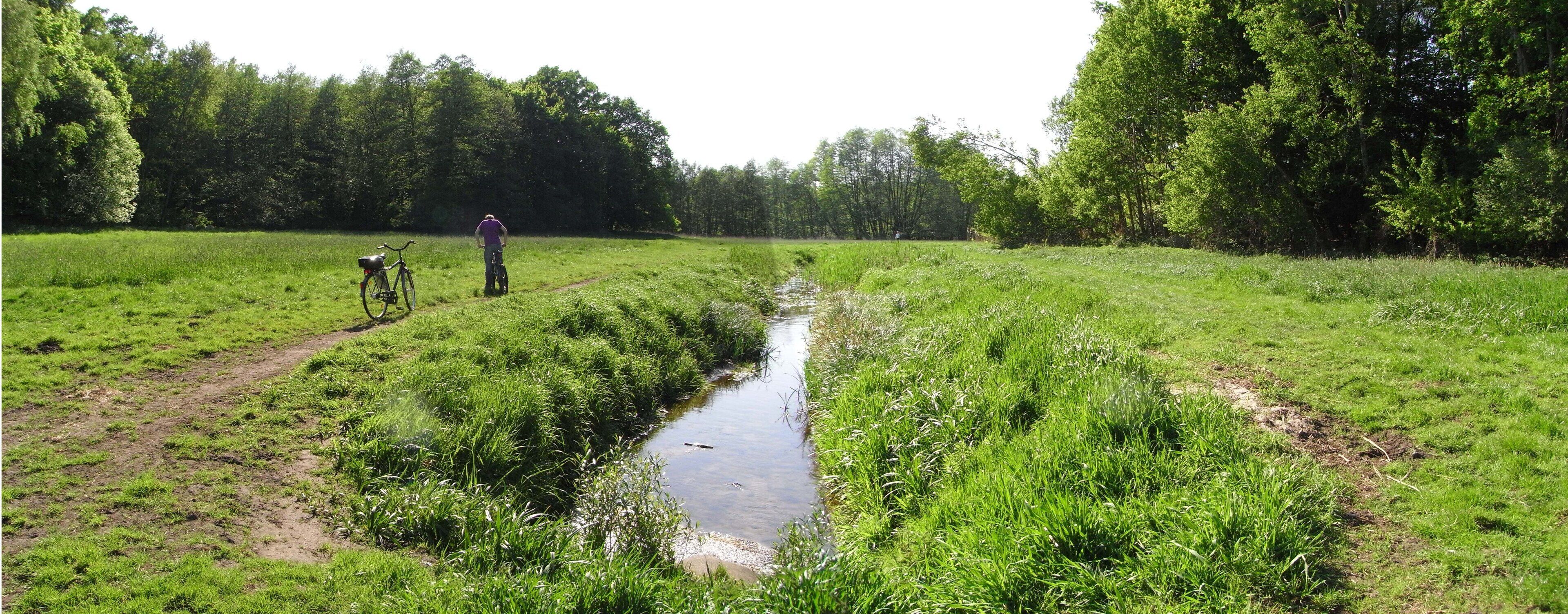 A narrow water channel in a grassy field with a bicycle and a person standing in the distance.