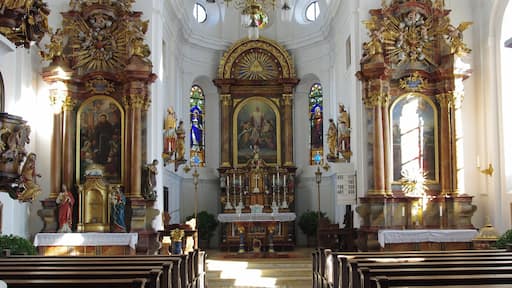 Interior de una iglesia ornamentada con un altar central, vidrieras de colores y filas de bancas.