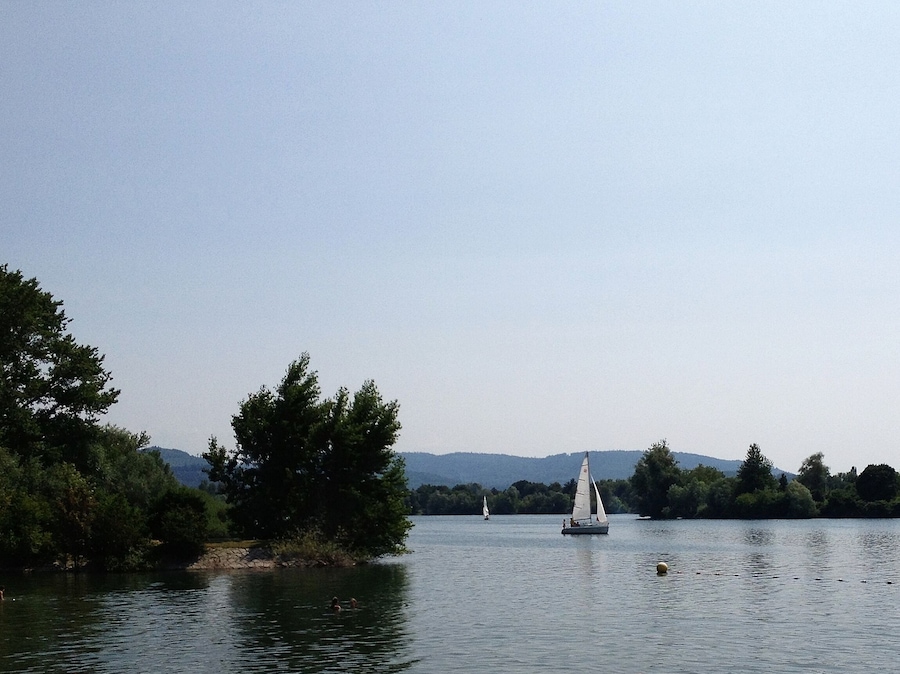A calm lake with a sailboat, surrounded by dense trees and distant hills.