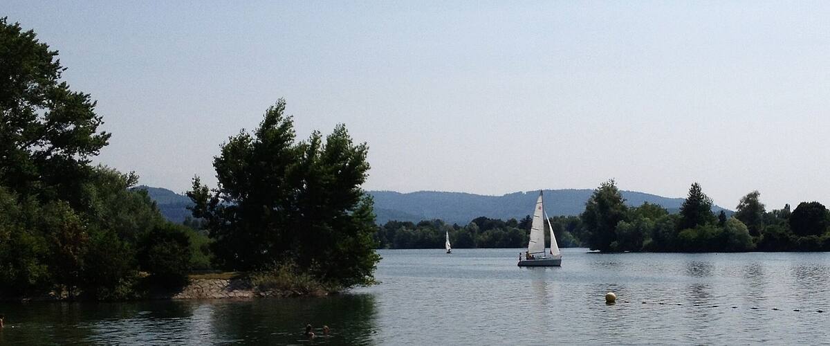 A calm lake with a sailboat, surrounded by dense trees and distant hills.