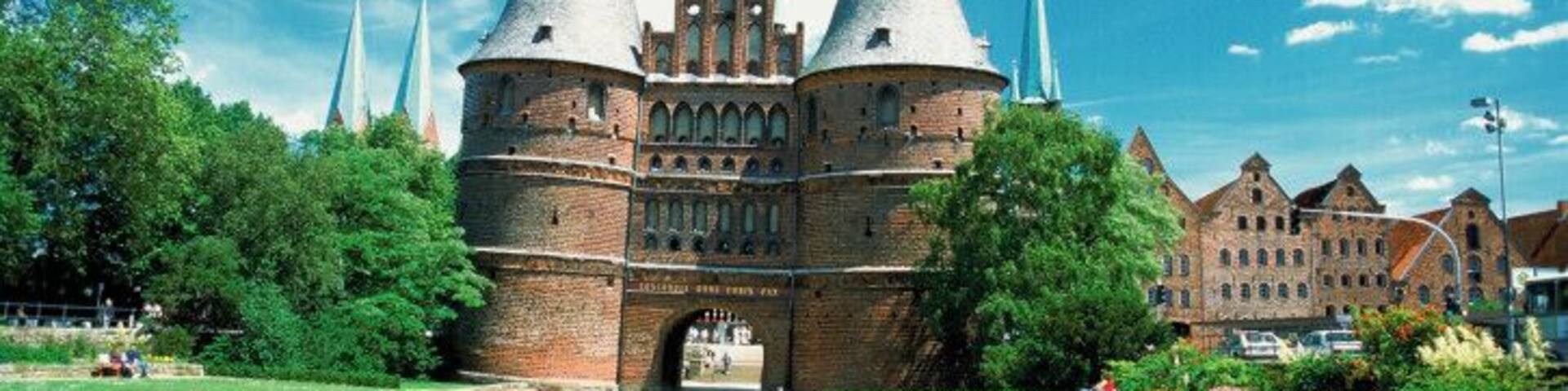 Un edificio histórico de ladrillo con dos torres puntiagudas y un gran arco, rodeado de un jardín cuidado con rosas rosadas.