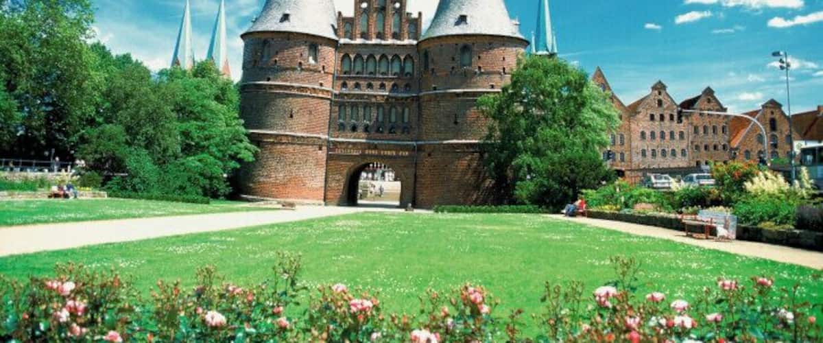 A historic brick building with two pointed towers and a large archway, surrounded by a well-maintained garden with pink roses.
