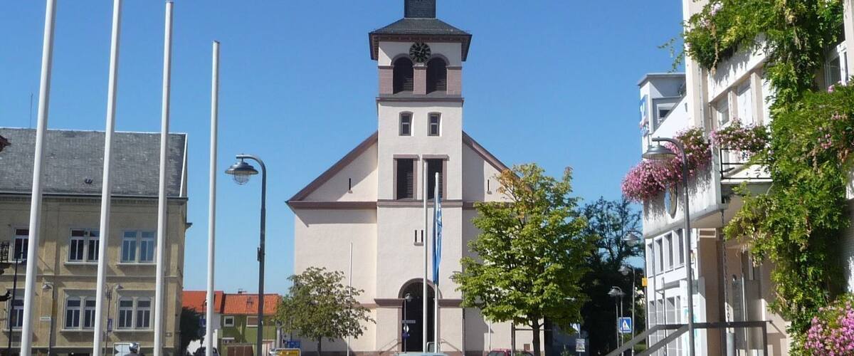A church with a clock tower, surrounded by a paved square with flower planters and modern buildings.