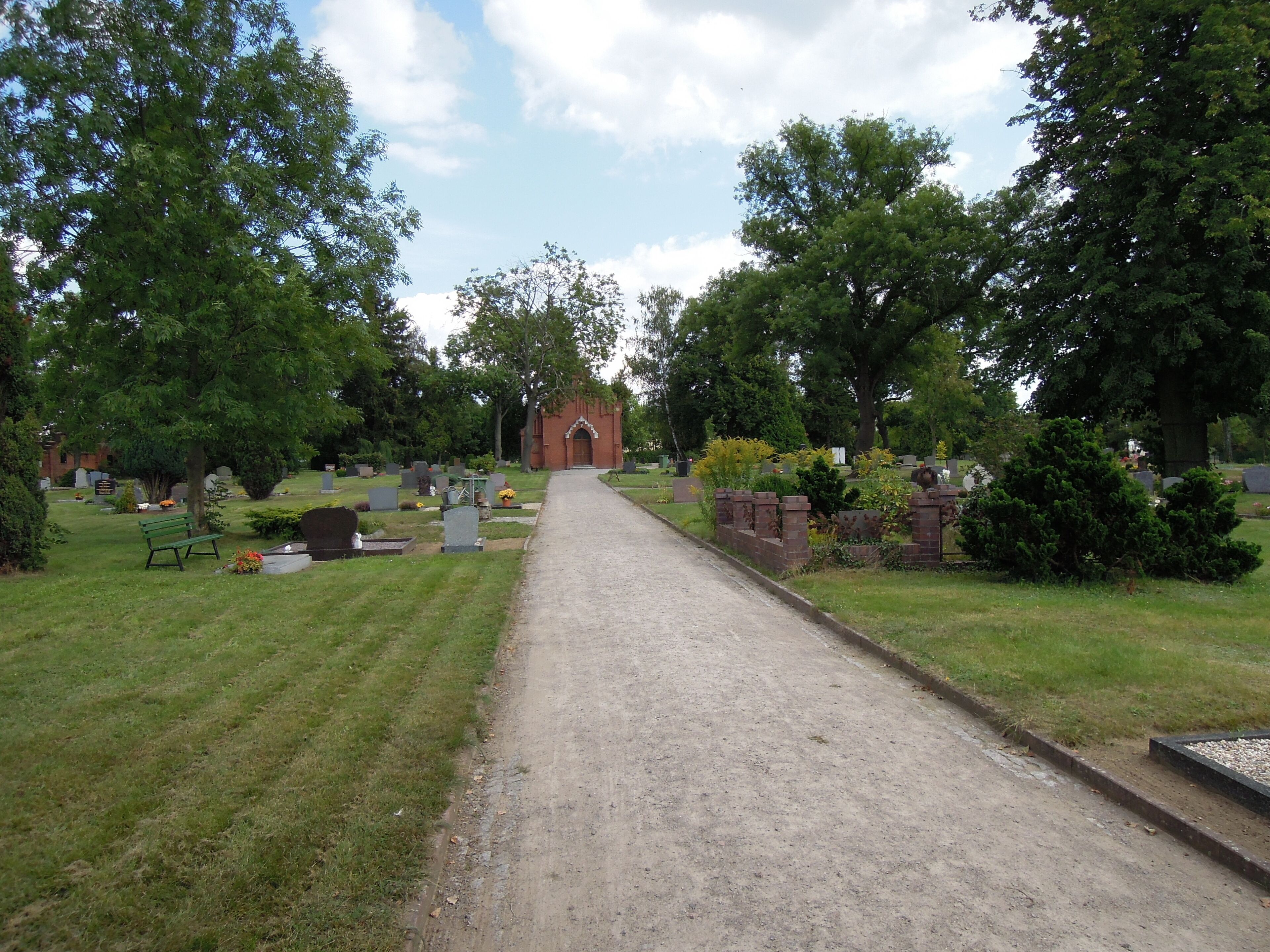 A cemetery with a gravel path leading to a brick building, surrounded by trees and headstones.