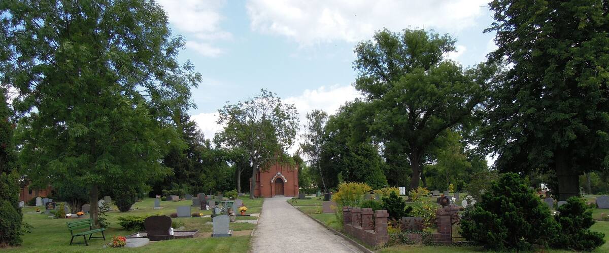 A cemetery with a gravel path leading to a brick building, surrounded by trees and headstones.