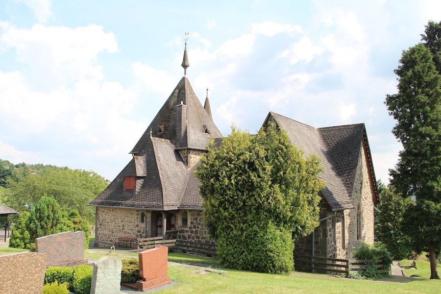 Une église en pierre, dotée d’un clocher en forme de flèche et d’un grand arbre devant.