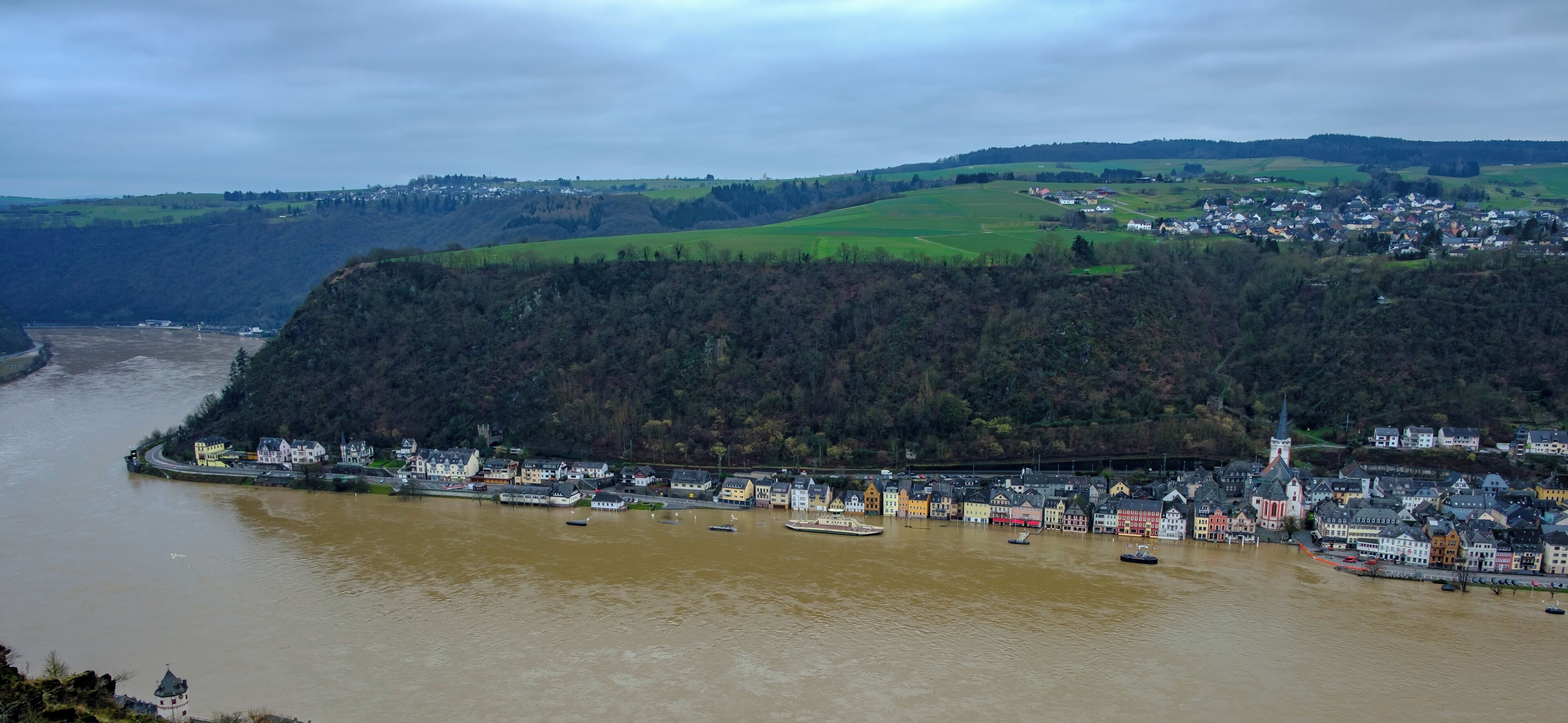 A river with boats, a town with colorful houses, and a forested hill.