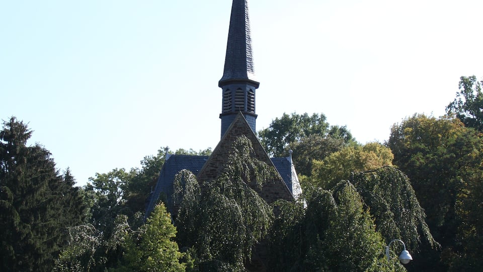 A church with a tall steeple and a cross at the top, surrounded by trees and a paved road.