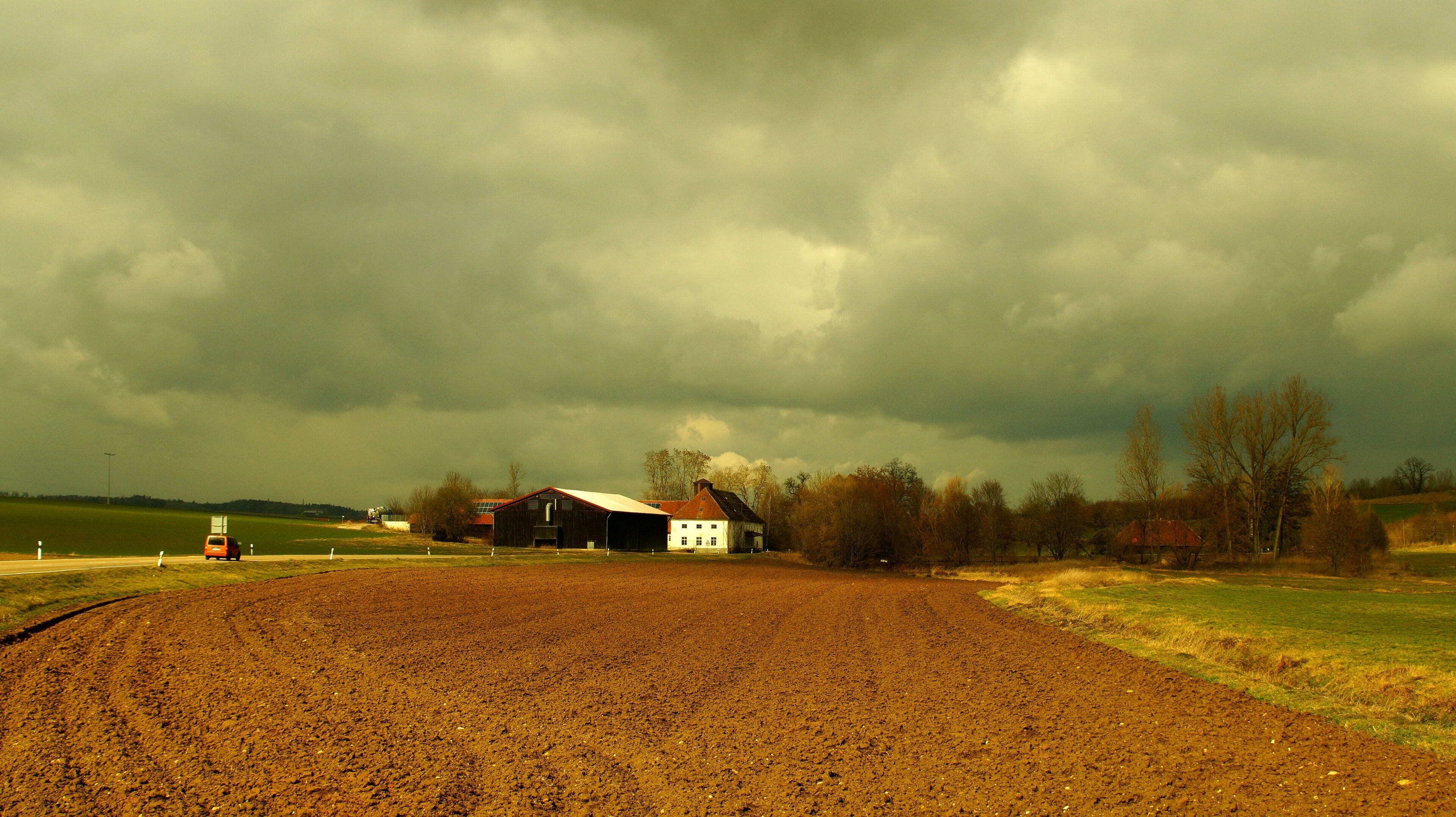 A rural landscape with a plowed field, a red car, a white barn, and a dark, cloudy sky.
