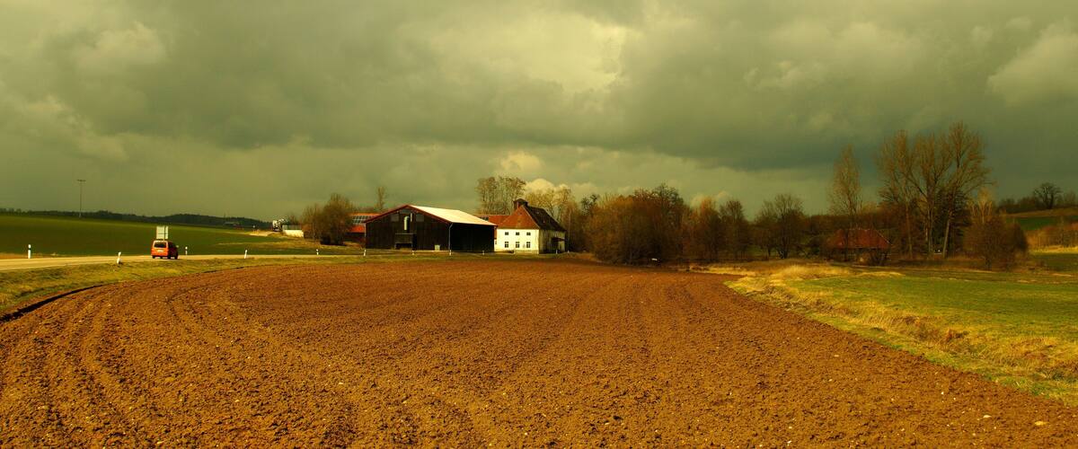 A rural landscape with a plowed field, a red car, a white barn, and a dark, cloudy sky.