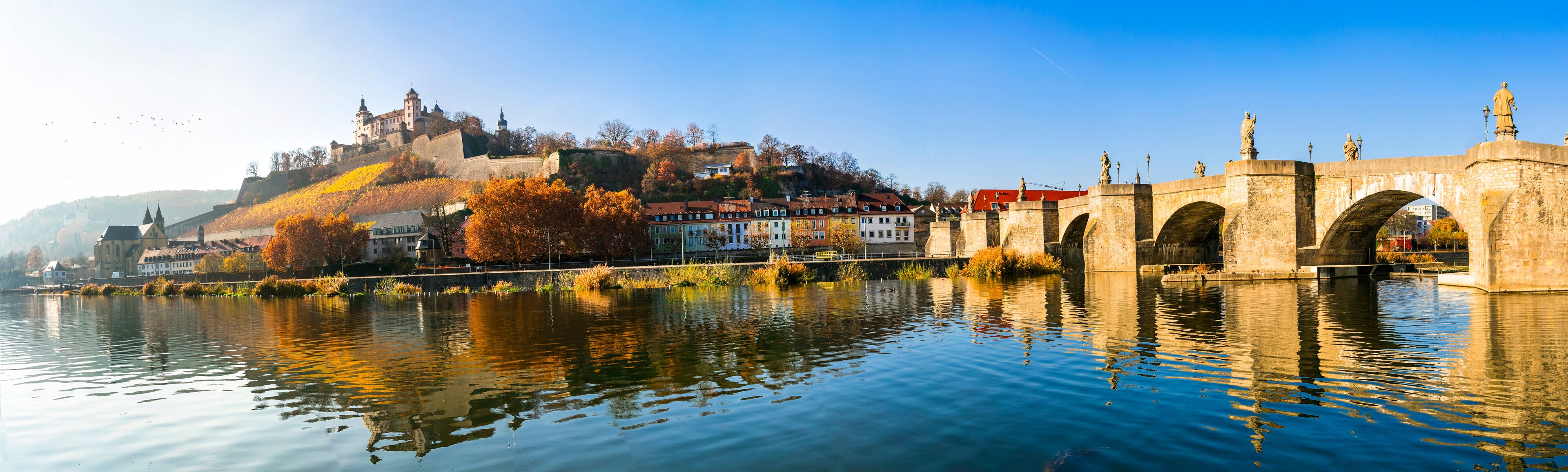 A river with a stone bridge, autumn-colored trees, and a castle on a hill.