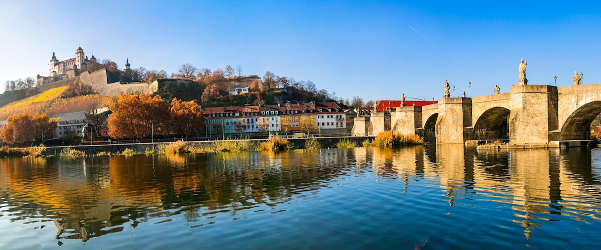 A river with a stone bridge, autumn-colored trees, and a castle on a hill.