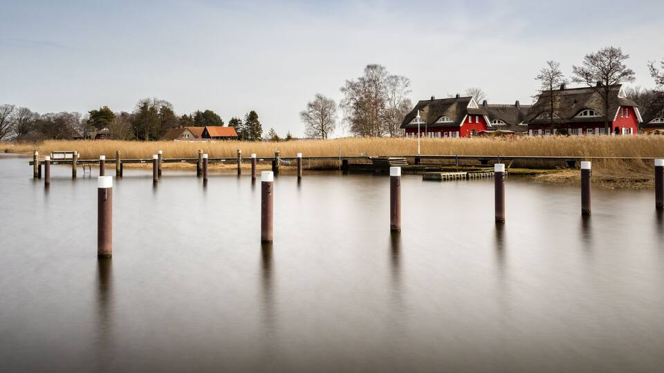 A calm lake with wooden posts in the foreground and traditional houses in the background.
