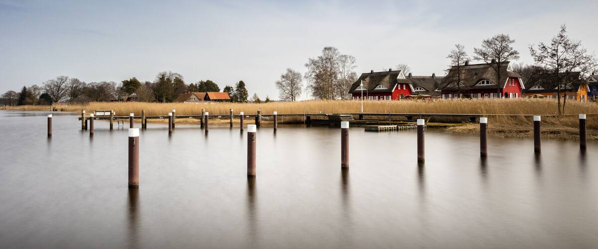 A calm lake with wooden posts in the foreground and traditional houses in the background.