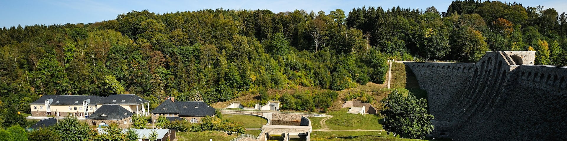 A stone dam with a water channel, surrounded by lush greenery and a forested hillside.