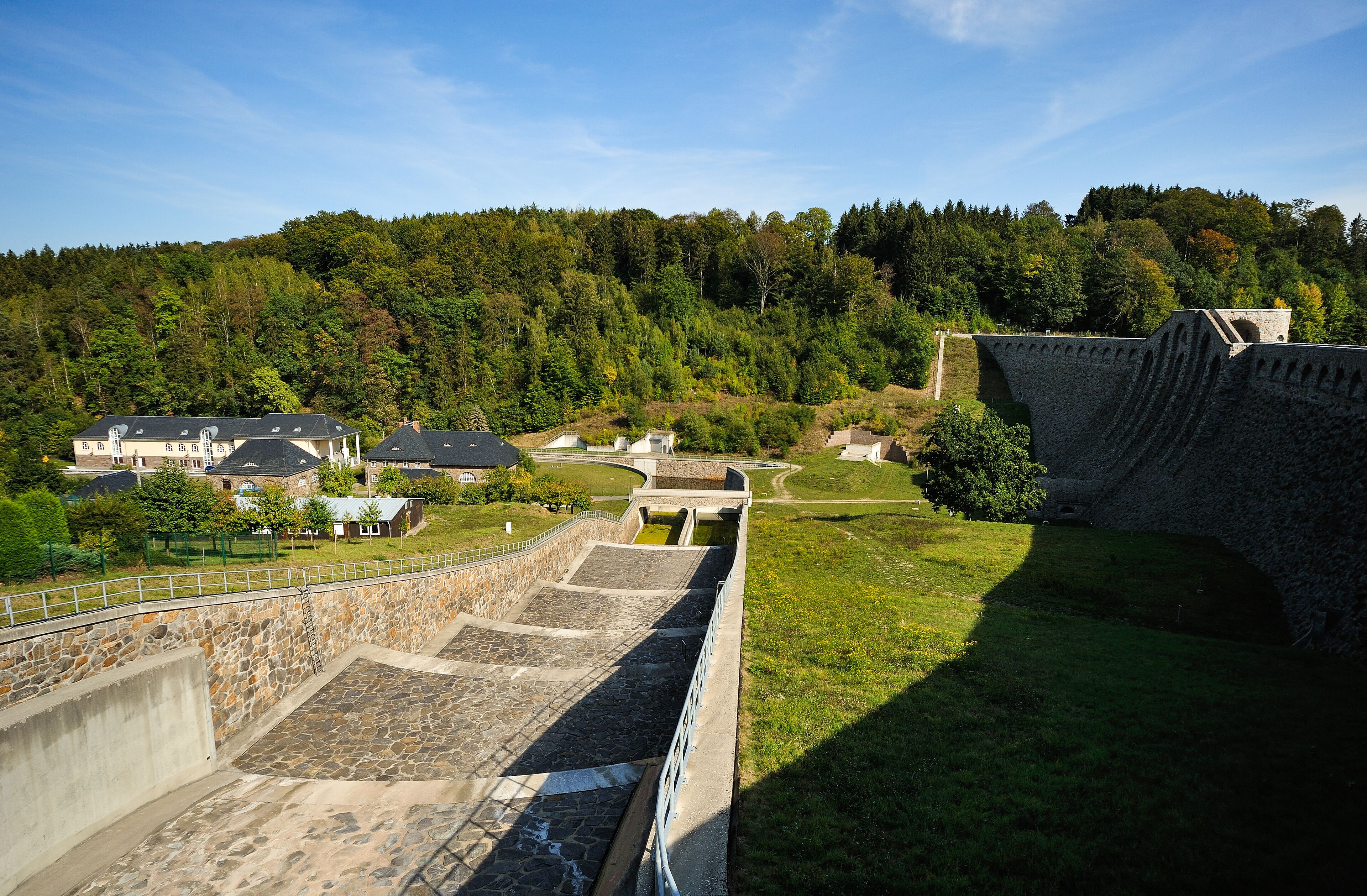 A stone dam with a water channel, surrounded by lush greenery and a forested hillside.