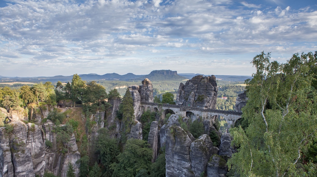 Paisagem com imponentes formações rochosas, uma ponte de pedra e uma cadeia montanhosa distante sob um céu parcialmente nublado.