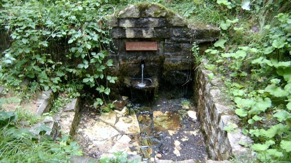 A rustic stone well with a sign reading "Rübersulzquelle" surrounded by lush greenery.