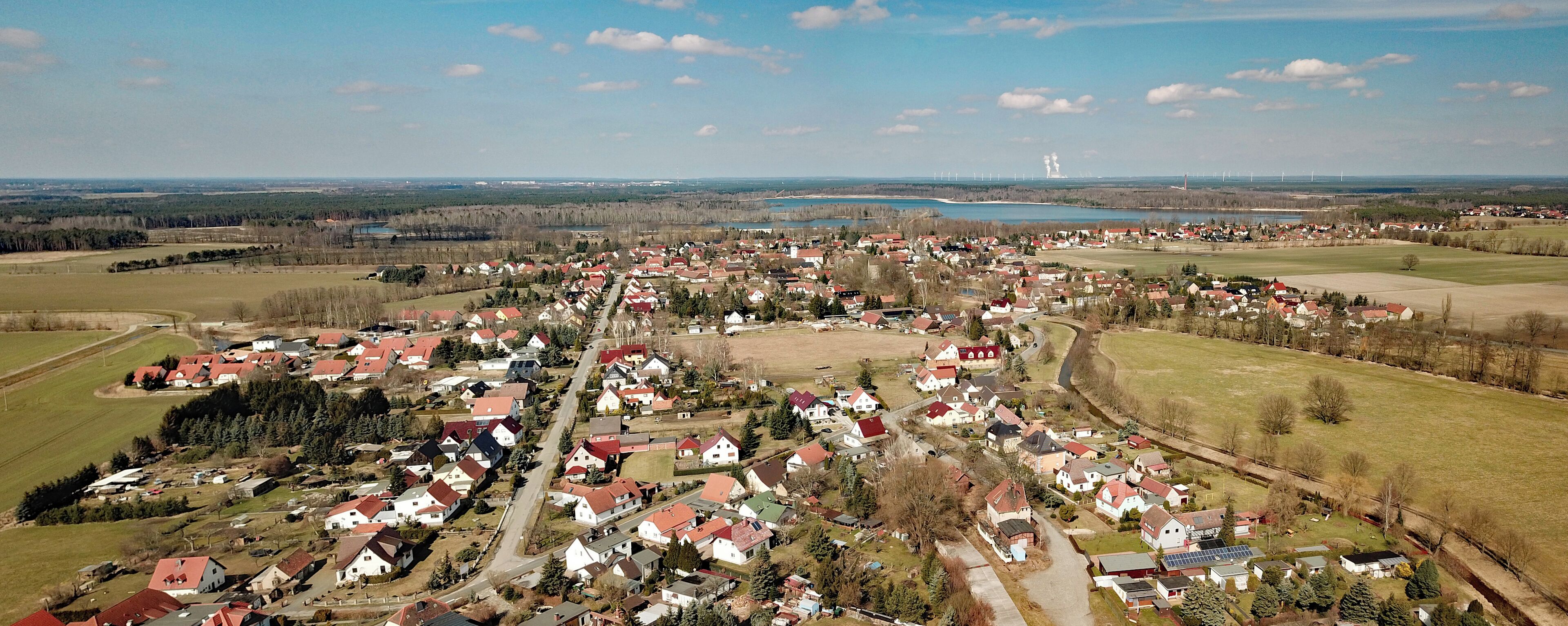 An aerial view of a rural village with houses, fields, a lake, and wind turbines in the distance.