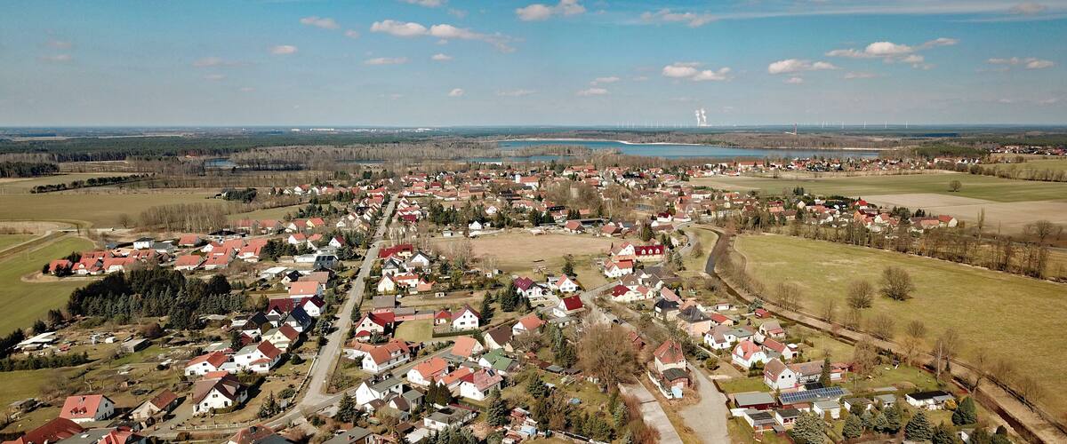 An aerial view of a rural village with houses, fields, a lake, and wind turbines in the distance.