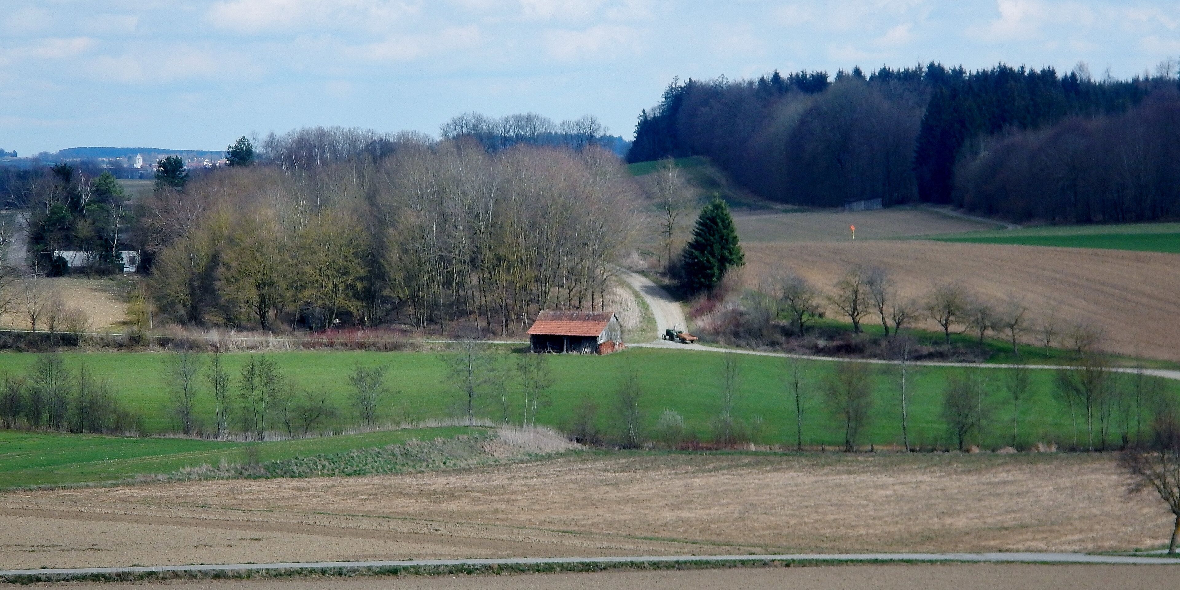 A rural landscape with a dirt road, a small red-roofed barn, and a tractor.