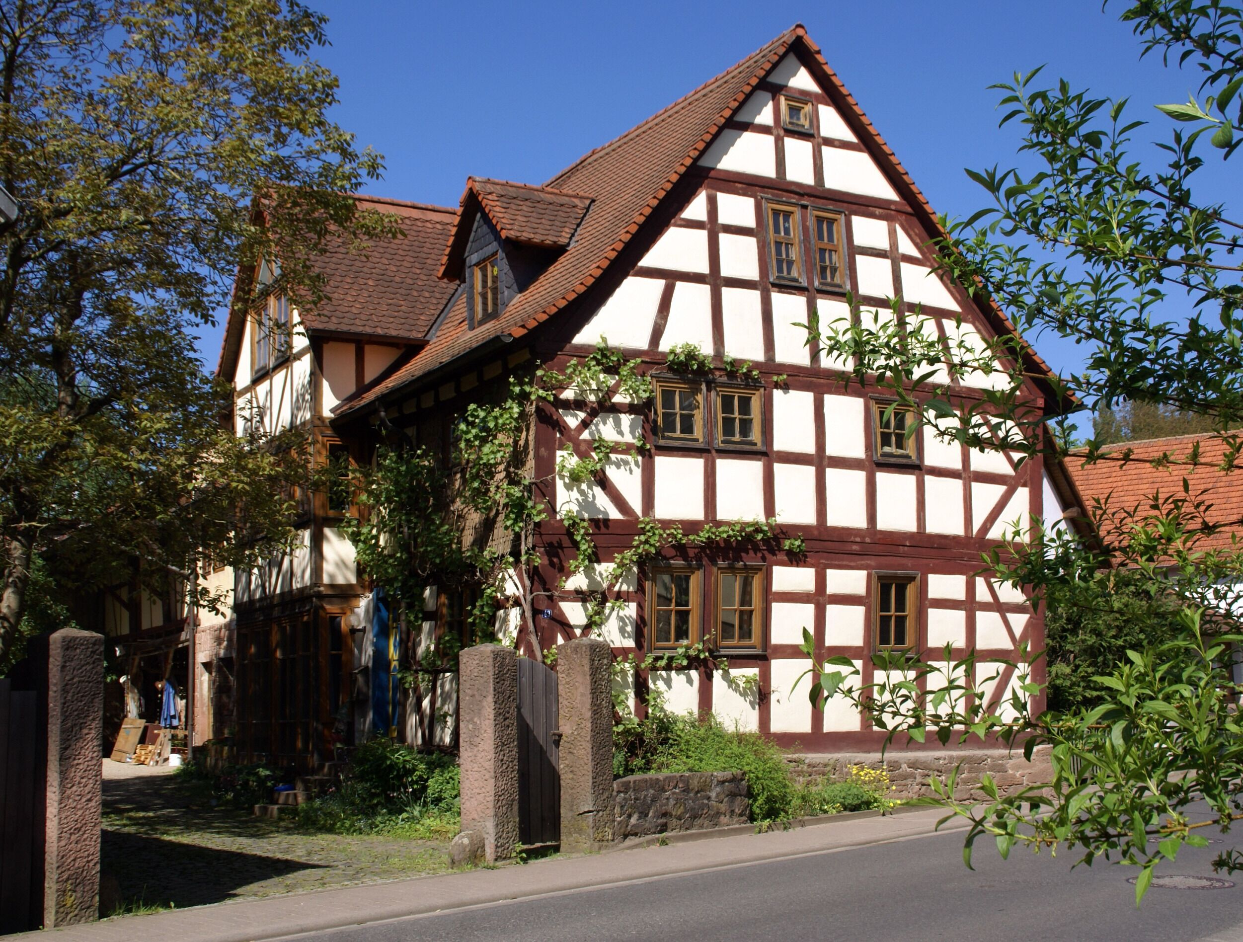 A traditional half-timbered house with a steeply pitched roof, surrounded by greenery and a cobblestone path.