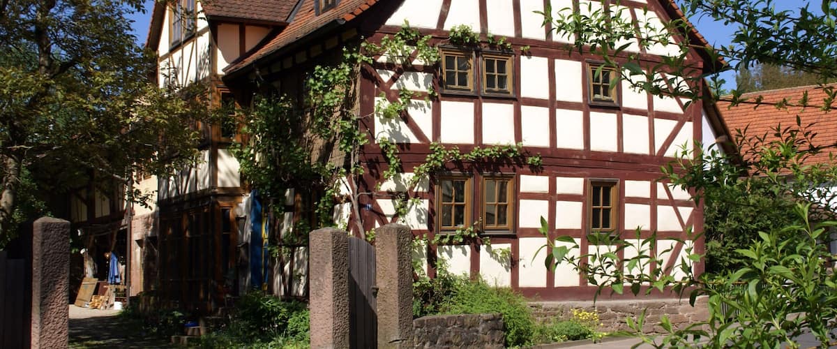 A traditional half-timbered house with a steeply pitched roof, surrounded by greenery and a cobblestone path.