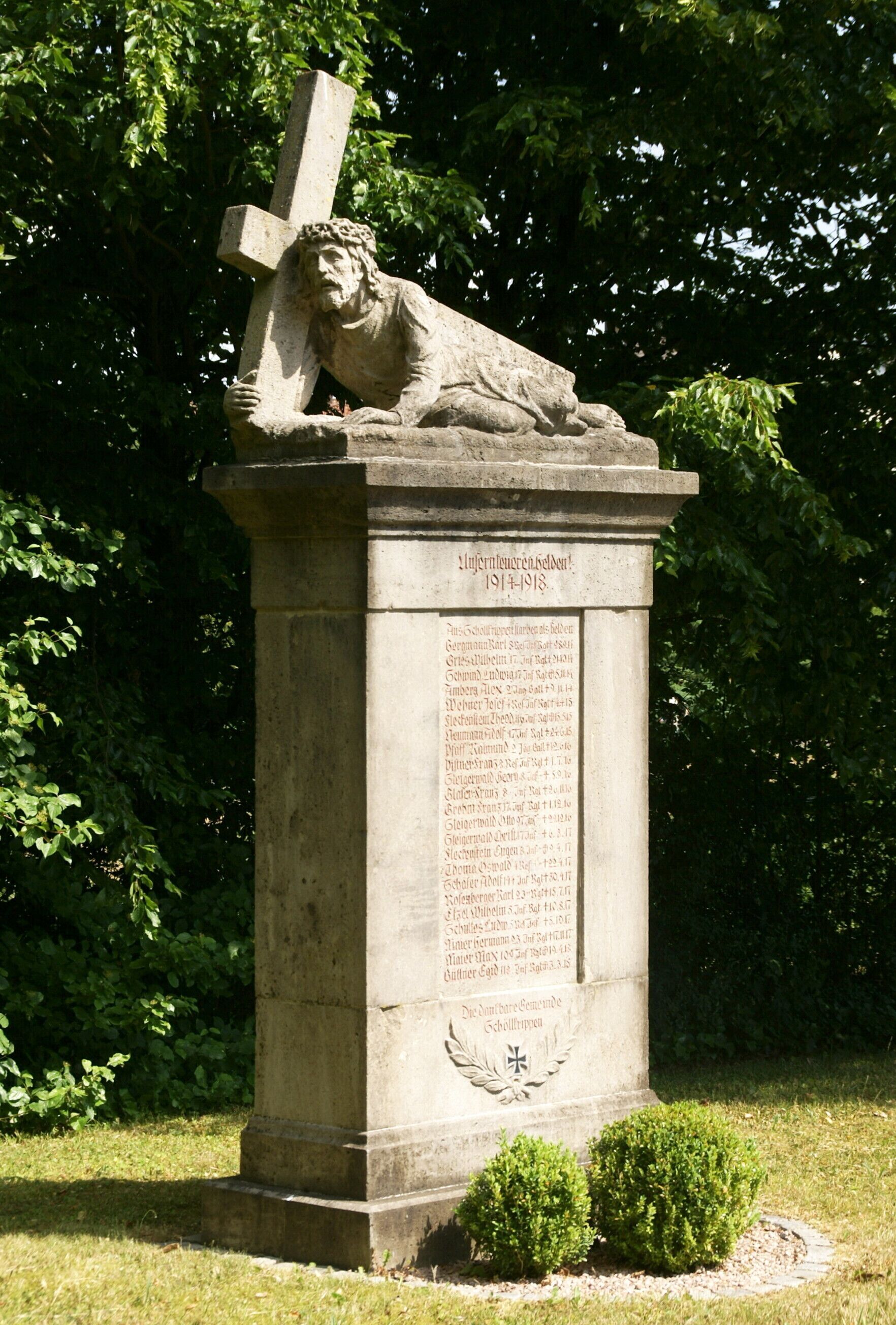 A stone monument with a cross and a figure lying beneath it, surrounded by greenery.
